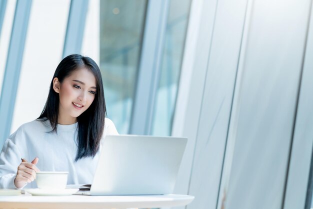 empresaria emprendedora dueña de una empresa atractiva mujer de negocios asiática comunicación con teléfono inteligente y computadora portátil vestido blanco alegre y sonrisa con confianza desenfoque de fondo de la oficina