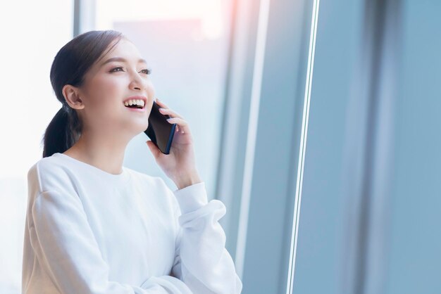 empresaria emprendedora dueña de una empresa atractiva mujer de negocios asiática comunicación con teléfono inteligente y computadora portátil vestido blanco alegre y sonrisa con confianza desenfoque de fondo de la oficina