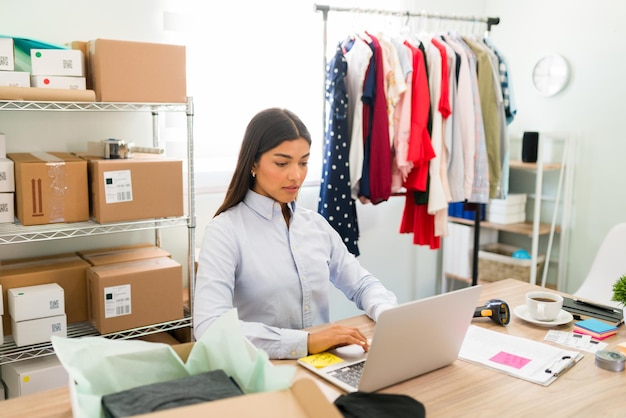 Empresaria enfocada sentada en su escritorio de oficina y actualizando los productos y la nueva ropa de moda en su comercio electrónico en línea