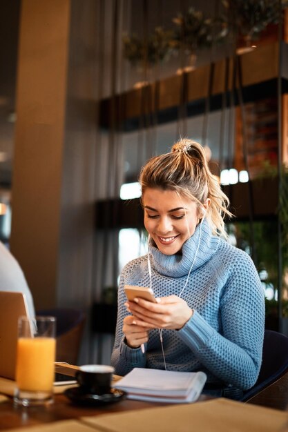 Empresaria feliz disfrutando de un descanso para tomar café mientras lee un mensaje de texto en un teléfono inteligente y escucha música con auriculares en un café