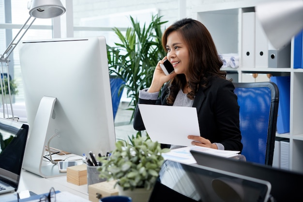 Empresaria filipina sonriente sentada en el escritorio en la oficina y hablando por teléfono móvil