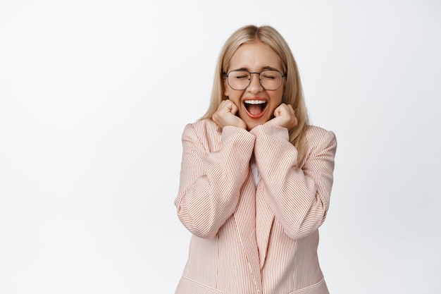 Empresaria gritando de asombro, reaccionando sorprendida y emocionada con una gran noticia, de pie en traje y gafas en blanco.