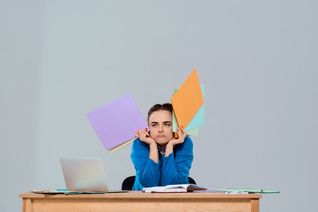 Empresaria hermosa joven cansada que se sienta en el lugar de trabajo, sosteniendo carpetas coloridas