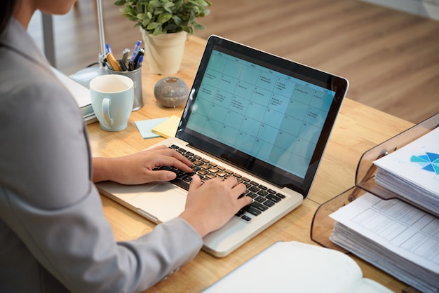 Empresaria irreconocible sentado en el escritorio con la computadora portátil y mirando el calendario