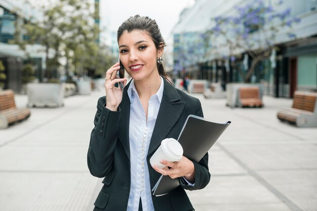 Empresaria joven acertada que habla en el teléfono celular que sostiene la taza y la carpeta de café disponibles en la mano