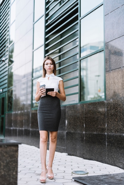 Empresaria joven confiada que se coloca delante del edificio de oficinas que sostiene la taza de café y el smartphone disponibles