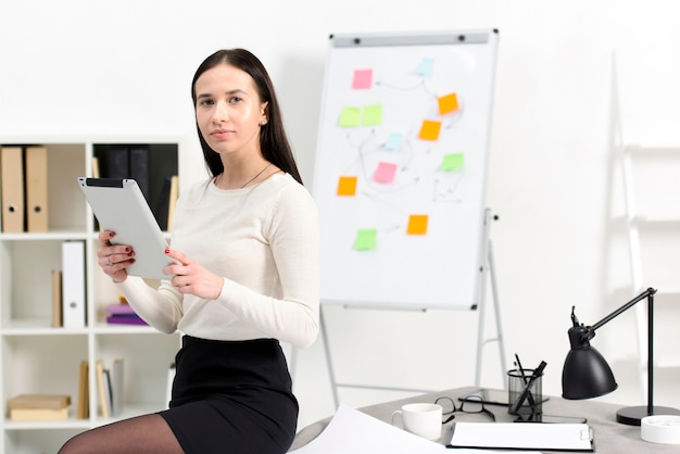 Foto gratuita empresaria joven confiada que sostiene la tableta digital en la mano que se sienta en la tabla en el lugar de trabajo