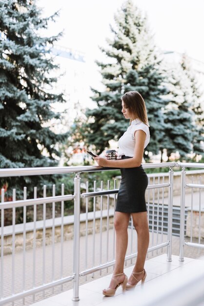 Empresaria joven elegante que se coloca en el balcón que mira el teléfono móvil