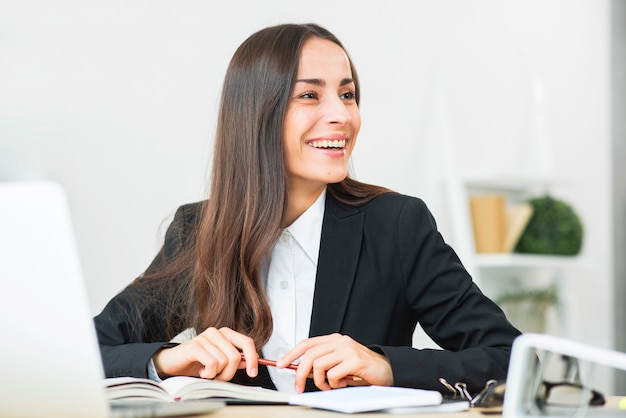 Empresaria joven feliz que se sienta en el escritorio de oficina que mira lejos