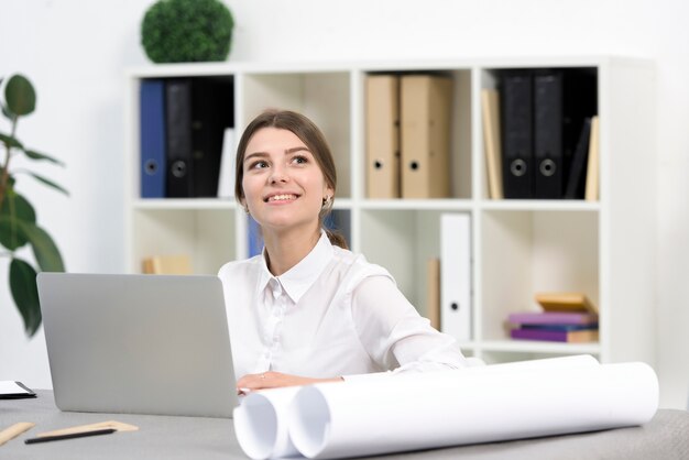 Empresaria joven feliz que sueña despierto mientras que usa el ordenador portátil en la oficina