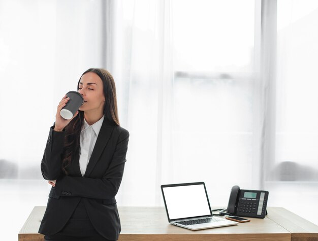 Empresaria joven que se coloca delante del café de consumición del escritorio de la taza disponible