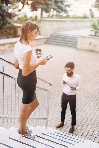 Empresaria joven que se coloca en la escalera usando smartphone mientras que hombre de negocios que mira la tableta digital