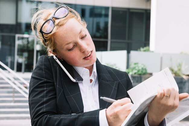 Empresaria joven que escucha en la escritura del teléfono móvil en el diario con la pluma