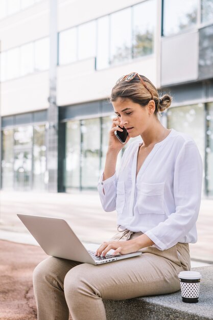 Empresaria joven que habla en el teléfono móvil usando la computadora portátil