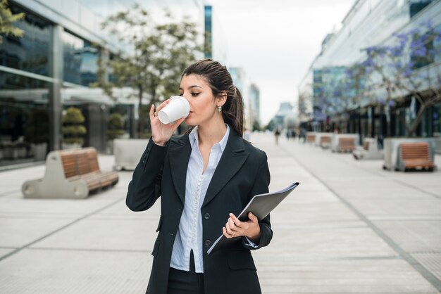 Empresaria joven que sostiene la carpeta a disposición que bebe el café en el campus del negocio