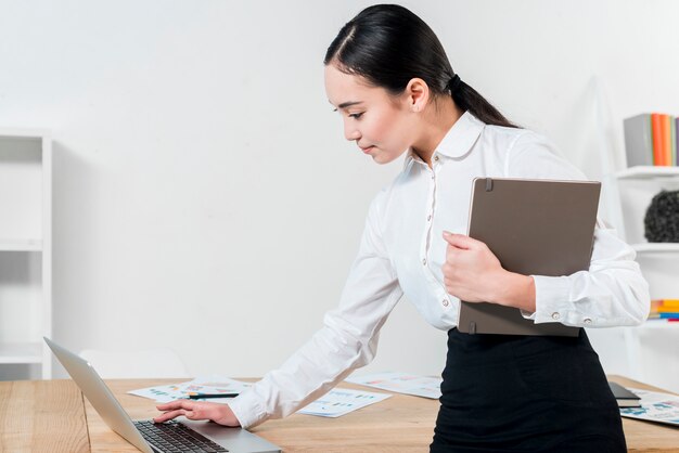 Empresaria joven que sostiene el diario a disposición usando el ordenador portátil en la tabla en el lugar de trabajo