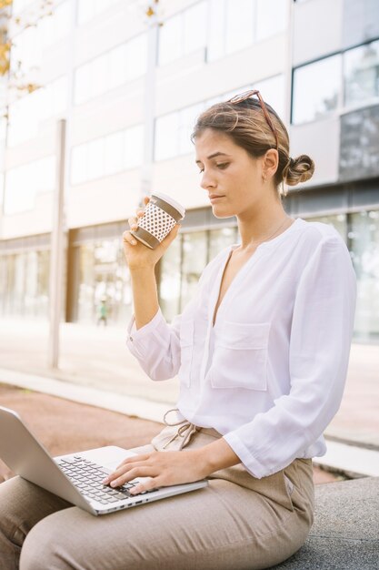 Empresaria joven que sostiene la taza de café para llevar que mecanografía en el ordenador portátil