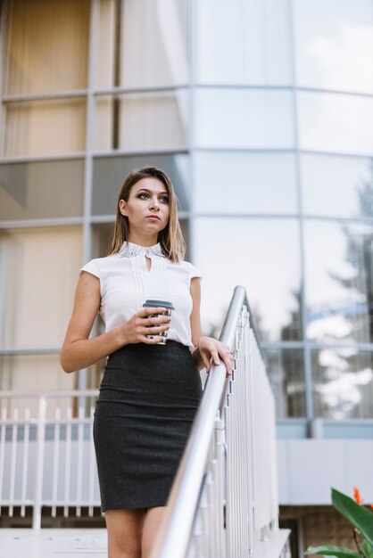 Empresaria joven rubia que sostiene la taza disponible que se coloca cerca de la verja delante del edificio de oficinas