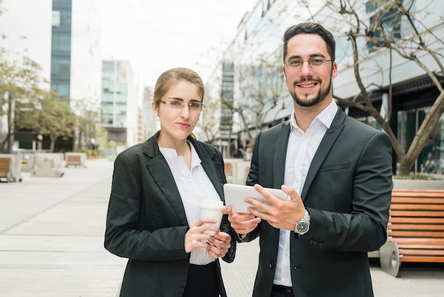 Empresaria joven sonriente y hombre de negocios que se colocan fuera de la oficina que sostiene la taza de café móvil y disponible