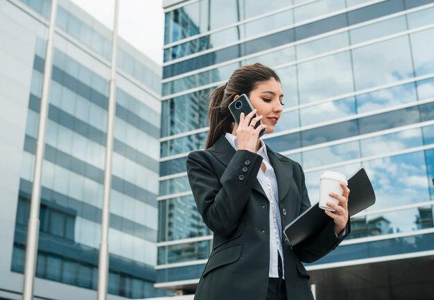 Empresaria joven sonriente que habla en el teléfono celular que se coloca delante del edificio