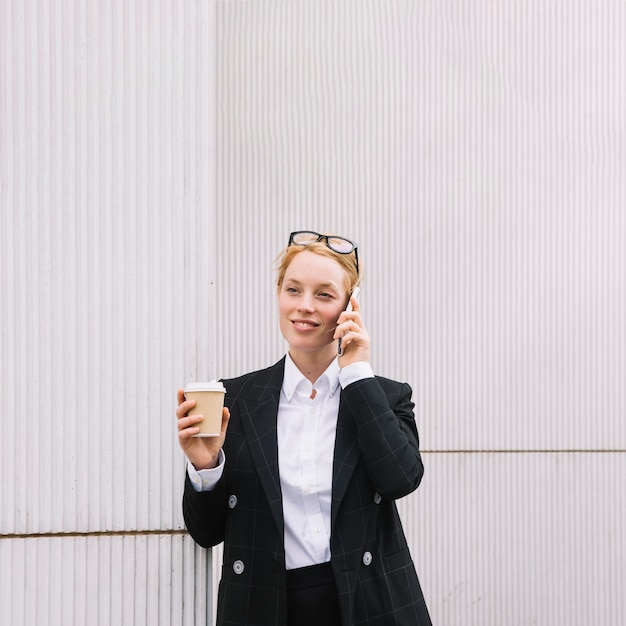 Foto gratuita empresaria joven sonriente que habla en el teléfono celular que sostiene la taza de café para llevar