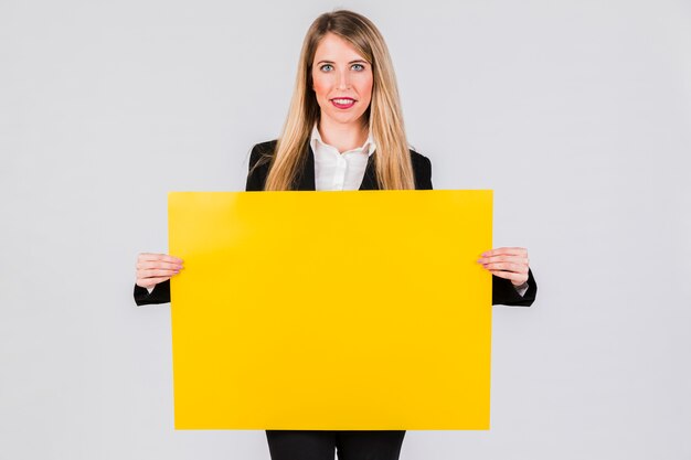 Empresaria joven sonriente que lleva a cabo el cartel en blanco amarillo contra fondo gris