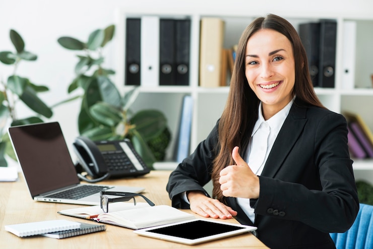 Foto gratuito empresaria joven sonriente que se sienta en el lugar de trabajo que muestra el pulgar encima de la muestra