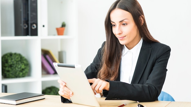 Empresaria joven sonriente que toca la tableta digital que se sienta en el escritorio de oficina