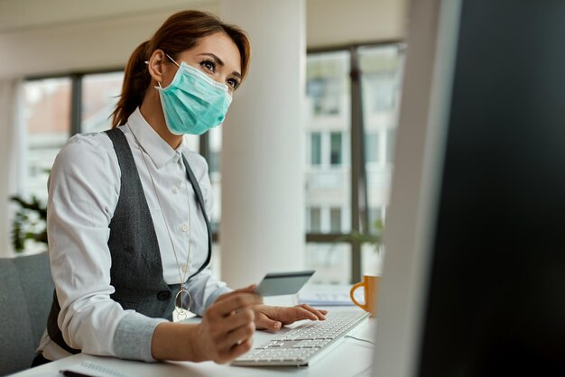 Empresaria con mascarilla usando computadora y comprando en internet con tarjeta de crédito en la oficina durante la pandemia de COVID19