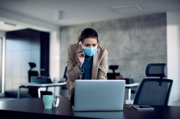 Foto gratuita empresaria con mascarilla usando una computadora portátil y comunicándose por teléfono móvil en su oficina