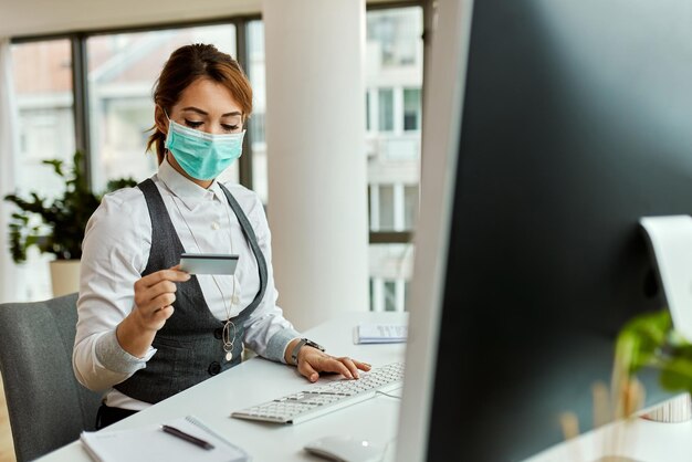 Empresaria con mascarilla usando computadora y tarjeta de crédito mientras compra en línea en la oficina