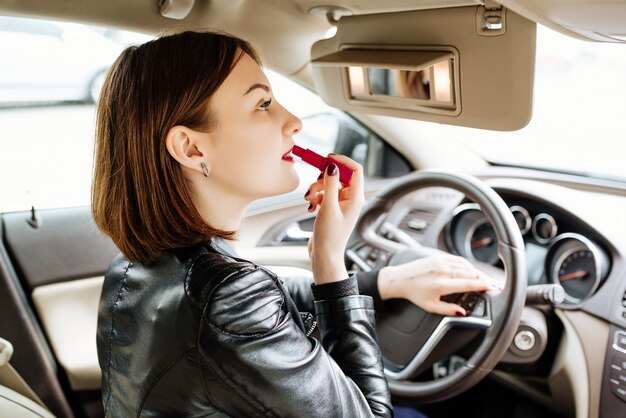 Empresaria mirando en el espejo retrovisor y haciendo sus labios con lápiz labial rojo en el coche