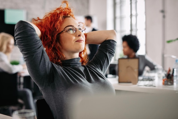 Foto gratuita empresaria pelirroja satisfecha descansando después del trabajo y soñando despierto en la oficina hay gente en el fondo