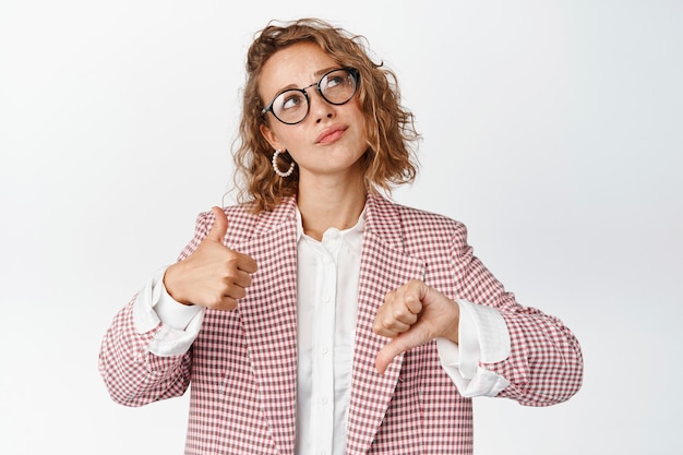 Empresaria pensativa con gafas y traje, mostrando los pulgares hacia arriba y hacia abajo, pensando en algo, tomando decisiones, sopesando los pros y los contras en blanco.