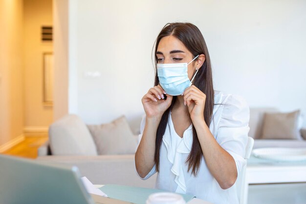 Empresaria poniéndose una máscara protectora trabajando en una computadora portátil en la oficina Trabajadora de empresa enfocada con máscara facial protectora trabajando en una computadora portátil en casa manteniendo las medidas de cuarentena del virus covid 19