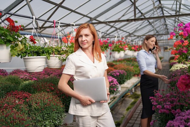 Empresaria posando con una computadora portátil mientras su pareja discutiendo por teléfono una propuesta en una casa verde con flores.