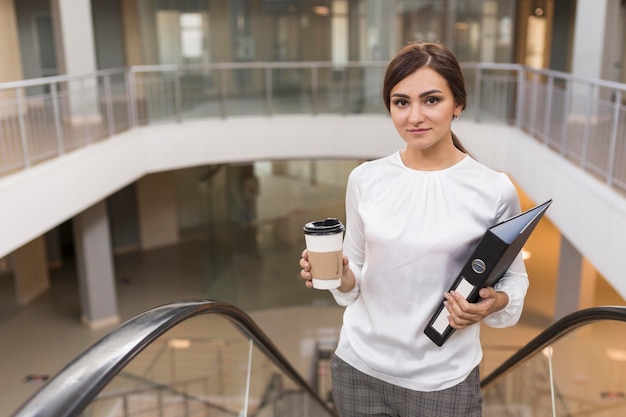 La empresaria posando en la escalera mecánica con café y carpeta