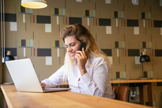 Empresaria positiva que trabaja en la computadora portátil y hablando por teléfono celular en el espacio de trabajo conjunto