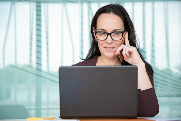 Empresaria preocupada en gafas hablando por teléfono móvil y abriendo los ojos, trabajando en la computadora en la oficina, usando la computadora portátil en la mesa