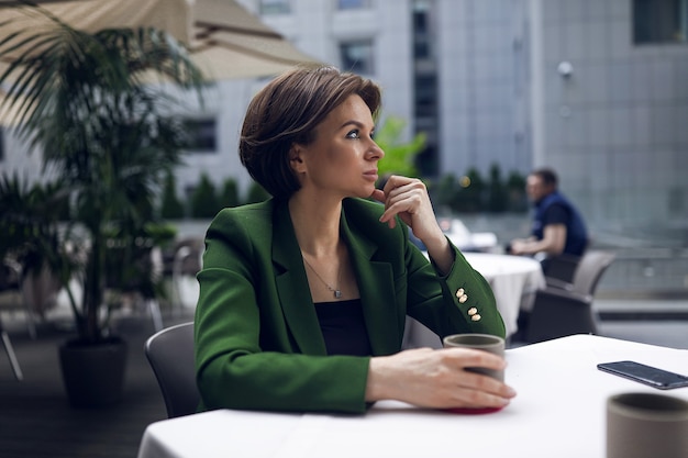 Empresaria sentada en la cafetería y descansar un poco después de todas las reuniones y entrevistas. Elegante chaqueta verde y blusa negra, corte de pelo corto, maquillaje nude. Taza de cafe caliente