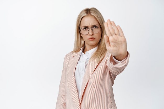 Empresaria seria extendiendo la mano en señal de stop, prohibir, prohibir algo, decir que no, de pie en blanco con traje y gafas