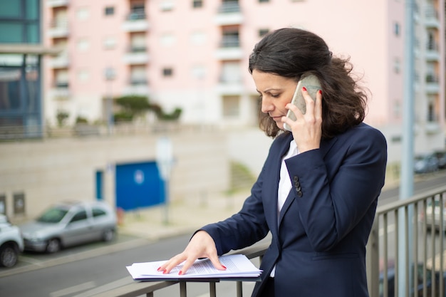 Foto gratuita empresaria seria hablando por teléfono inteligente y mirando papeles