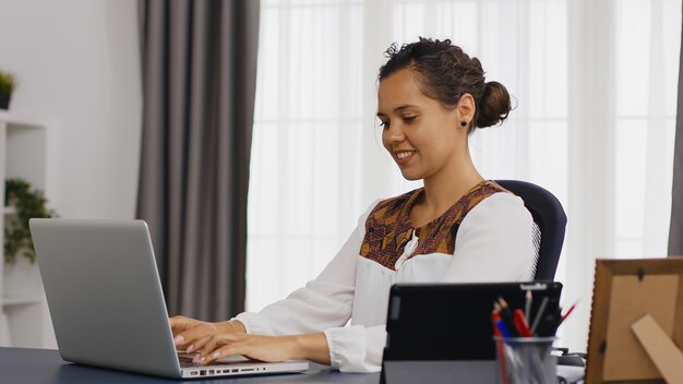 Empresaria sonriente escribiendo en la computadora portátil desde la oficina en casa.