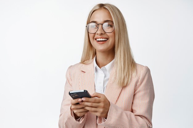 Empresaria sonriente en gafas y traje, mediante teléfono móvil, de pie en el estudio blanco