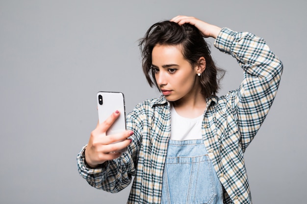 Empresaria sonriente haciendo foto selfie en smartphone. Vistiendo con camisa azul y gafas. De pie sobre una pared gris