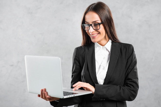 Empresaria sonriente que mira la computadora portátil en su mano contra el muro de cemento