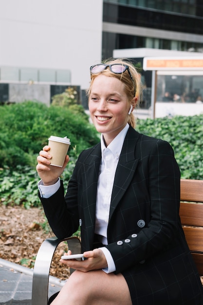 Foto gratuita empresaria sonriente que se sienta en el banco que sostiene el teléfono móvil y la taza de café disponible