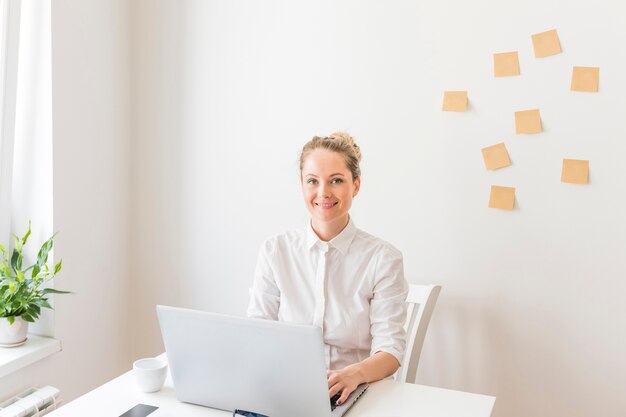 Empresaria sonriente que usa la computadora portátil con notas adhesivas en la pared