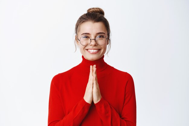 Empresaria sonriente en vasos tomados de la mano en gesto de namaste, agradecida y complacida con la ayuda, de pie contra la pared blanca