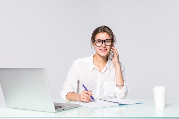 Empresaria en su escritorio de trabajo con ordenador portátil y teléfono parlante aislado sobre fondo blanco.